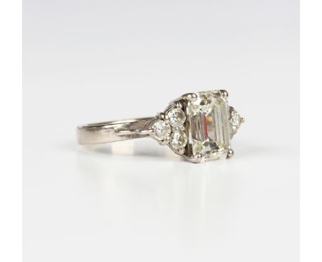 A white gold and diamond ring, claw set with the principal emerald cut diamond between circular cut diamond set three stone s