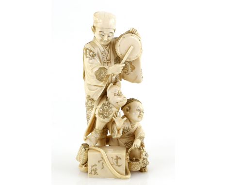 A stained ivory okimono carved as a standing entertainer beating a drum and balancing a kitsune mask on his left knee; a boy 