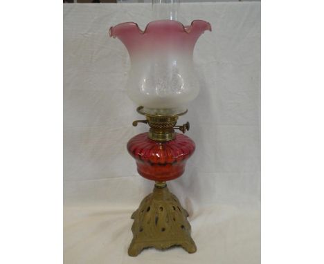 A Victorian oil lamp with cast iron base, cranberry tinted glass reservoir and shade with brass mounts