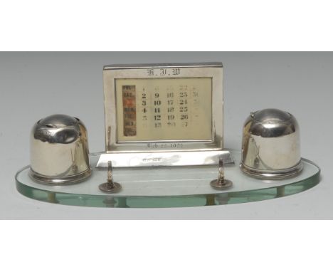 An Art Deco silver and clear glass oval desk stand, the rectangular calendar and pen rests flanked by a pair of domed inkwell