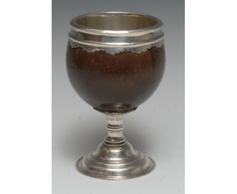 A George III silver coloured metal lined and mounted pedestal coconut cup, shaped pendant border, knopped stem, domed foot, 1