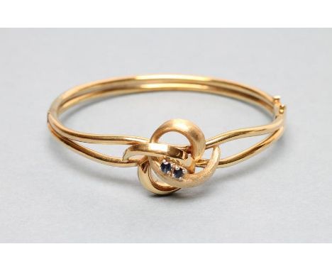A 9CT GOLD STIFF HINGED BANGLE, the open upper section centred by a knot claw set with two small sapphires, London 1976 (Est.