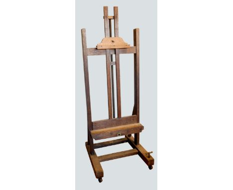 An early 20th century oak studio easel, the 'H' frame with bevel gear mechanism, with shelf and adjustable upper support on t