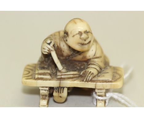 A Japanese carved ivory netsuke, 19th century, of a squinting man kneeling at a table with a brush in his hand, signed, 3.5cm