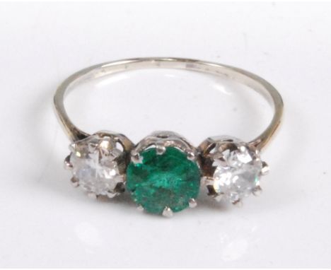 A white metal, emerald and diamond three-stone ring, comprising a centre round faceted emerald flanked to either side by a ro