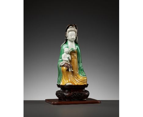 A BISCUIT PORCELAIN FIGURE OF GUANYIN WITH CHILD, KANGXI PERIODChina, 1662-1722. The goddess of mercy modeled seated at ease 