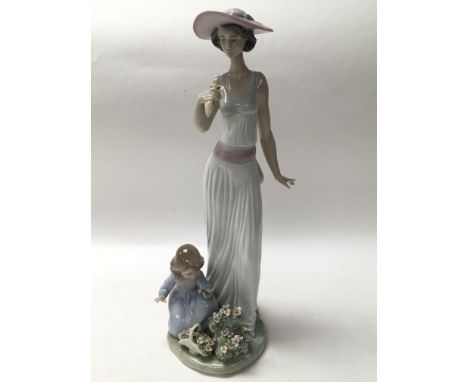 A Lladro figure of a lady wearing a hat, accompanied by a child who is picking flowers. The height is approximately 37cm. Wit