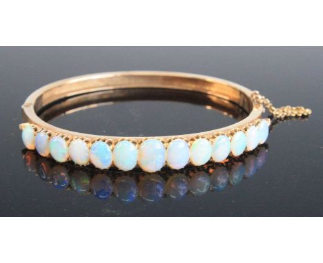 A rose metal opal oval hinged bangle, with box clasp and safety chain, featuring 13 graduated oval opal cabochons in claw set