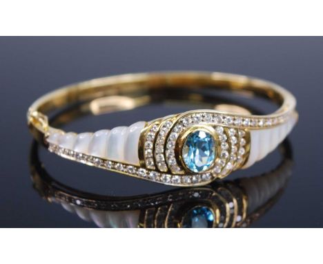 A yellow metal blue topaz, diamond and mother of pearl hinged oval bangle, featuring a centre oval blue topaz in a bezel sett