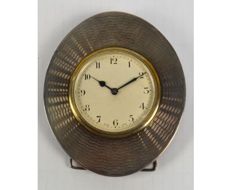 An Edwardian white metal framed oval time piece with overall engine-turned decoration, the circular enamel dial set with Arab