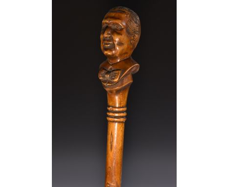 An early 20th century novelty softwood walking stick, the oversized pommel boldly carved with the bust of a gentleman, wearin