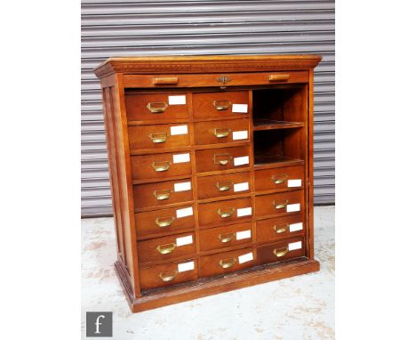 A 1920s oak filing cabinet fitted with an arrangement of eighteen drawers and a shelf, brass slot cup handles enclosed by a t