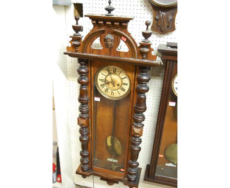 A late 19th century Vienna walnut drop trunk wall clock, having a turned and mask carved top, enamelled circular dial, twin w