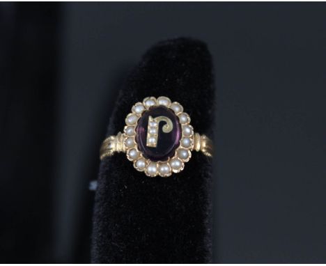 A yellow metal ring set with central purple stone inlaid with initial J, set with seed pearls and seed pearl border, ring siz