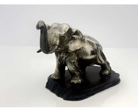 A white metal model of a strolling elephant with trunk raised, mounted on a wooden plinth, 13cm high, 21cm wide