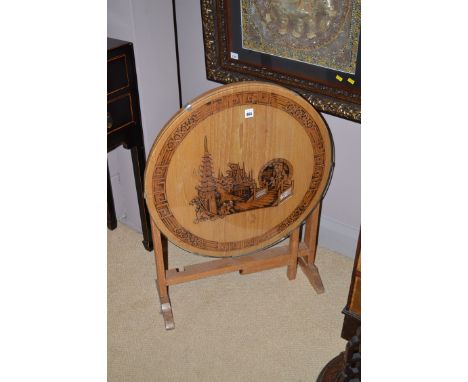 A carved light stained wood Chinese style tip-up-top occasional table, decorated with figures and pagodas in a landscape, wit