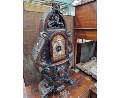 A MANTEL CLOCK IN AN OAK CASE CARVED WITH FOLIAGE ABOUT A CHILDS MASK BELOW THE SILVERED DIAL WITH ARABIC NUMERALS