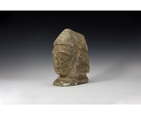 An undated archaistic limestone protome with gabled headdress, facial finial with fierce expression and bands of hair to the 