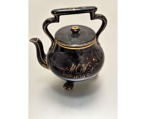 A Victorian black glazed pottery teapot highlighted with gold borders with gilded initials MEW 1875, raised on three paw feet