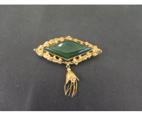 A jade brooch - The kite-shape nephrite jade panel, within a textured surround, suspending a foliate tassel - Tests as higher