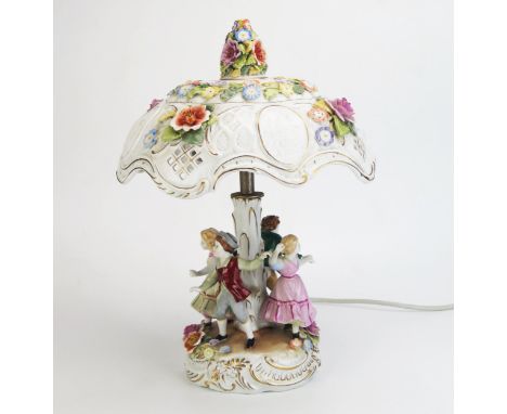 A German porcelain table lamp, with pieced domed shade raised on a stem with four children dancing below, on a shaped base wi