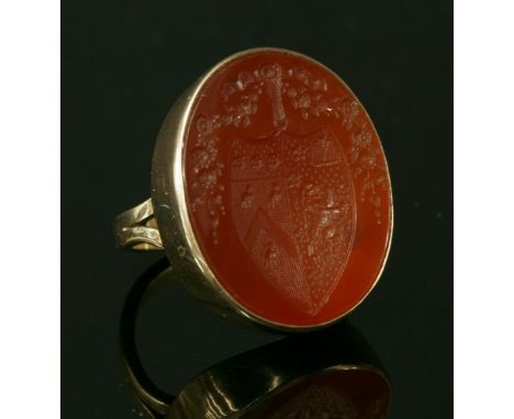 A gold intaglio carved cornelian ring,with an oval cabochon tablet intaglio entwined with a coat of arms, rub set to a plain 