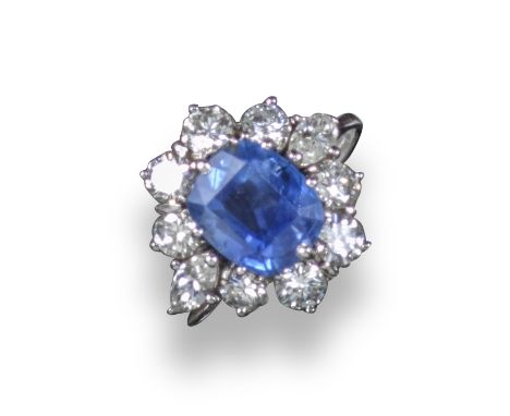 A sapphire and diamond cluster ring, the cushion-shaped sapphire is set within a surround of round brilliant-cut diamonds and