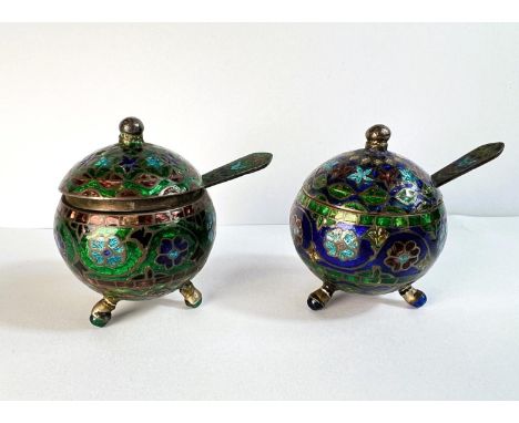 A pair of white metal lidded cruets with spoons, spherical, on ball feet, with colourful enamel decoration.