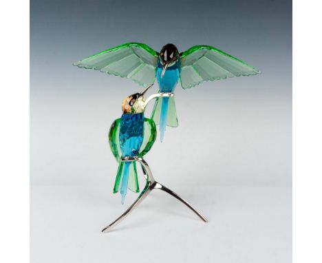 A stunning figurine of two birds made with green, blue, and amber colored crystal resting on a rhodium stand. Swarovski backs