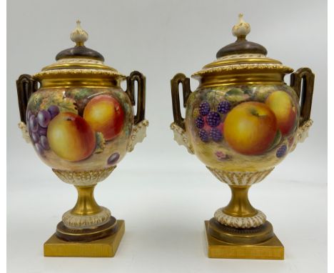 A pair of Royal Worcester twin handled lidded urns with masks to sides on a circular foot with a square plinth decorated with