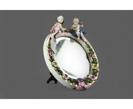 LATE 19TH CENTURY CONTINENTAL PORCELAIN MIRROR, of oval form, the frame surmounted with cherubs and applied with foliage, wit