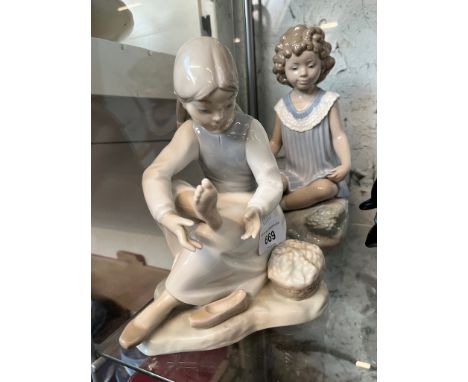 Two Nao figurines. seated girl in pleated dress, and seated girl looking at her foot with shoe removed 