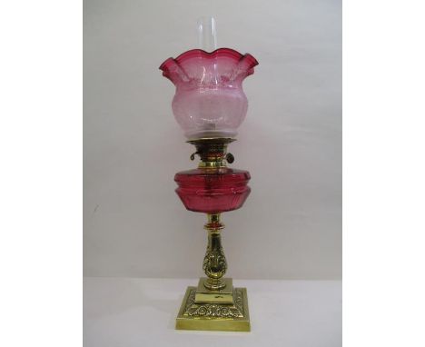 A Victorian brass oil lamp with an etched cranberry and clear glass shade and a cranberry glass reservoir, on an acanthus emb