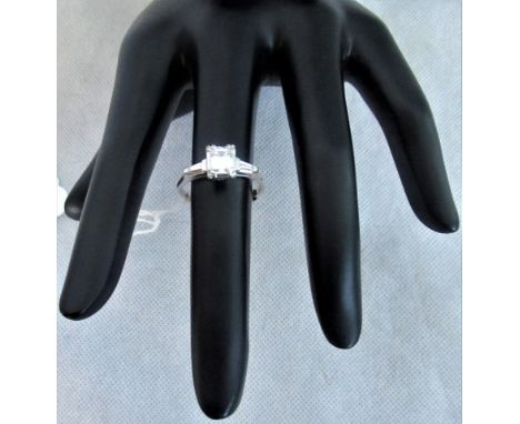 A single stone baguette cut diamond ring with simple four claw mounts, having trapeze cut diamonds to each shoulder and plain