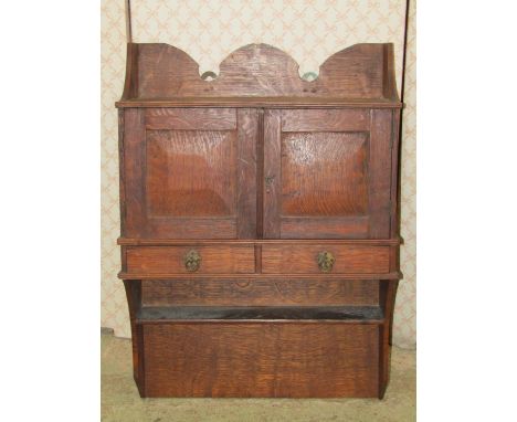 A small Arts &amp; Crafts style oak hanging wall cupboard with shaped outline and enclosed by a pair of panelled doors over t