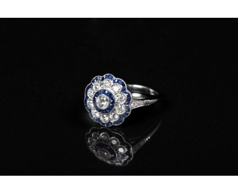 Diamond and sapphire flower-head cluster ring with central brilliant cut diamond surrounded by calibre cut blue sapphires, te