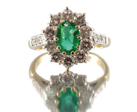 An 18ct gold oval-shape emerald and brilliant-cut diamond cluster ring, with diamond line shoulders, emerald estimated weight