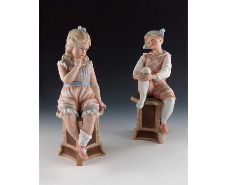 A pair of Gebruder Heubach bisque figures of  children, both modelled sitting on octagonal topped tables, the girl crying, th