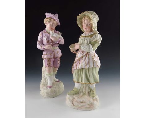 A pair of Gebruder Heubach bisque figures of children, both in pink and green costumes, gilt encrusted, modelled standing, th