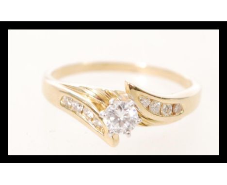 A ladies gold serpentine ring prong set with a brilliant cut diamond with further diamond accent stones to the shoulders. Dia