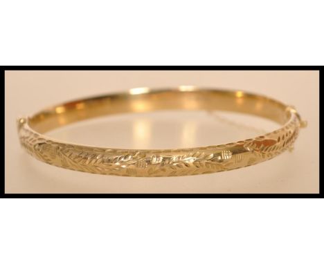 A hallmarked 9ct gold bangle with a hinged opening and safety chain with floral engraving. Hallmarked Sheffield. Approx 6cm i