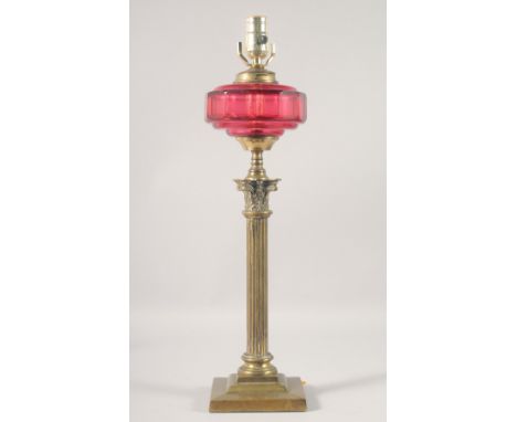 A BRASS CORINTHIAN COLUMN OIL LAMP with cranberry glass reservoir, (converted to electricity). 22ins high.