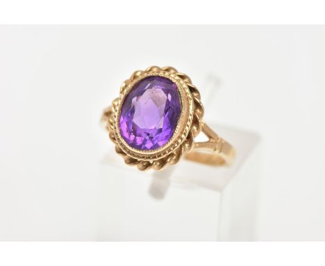 A 9CT GOLD AMETHYST DRESS RING, an oval cut amethyst set in a yellow gold bezel setting and a rope twist surround, leading on