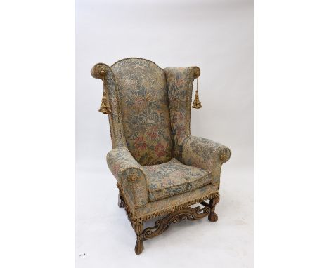 A high wing back armchair, in the 18th Century style, having tapestry upholstery, raised on carved front supports united by a