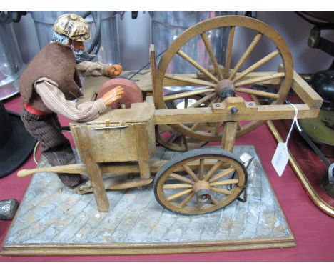 A Mid XX Century Electrically Operated Continental Automaton of a Knife Grinder, at work on his portable barrow with attendan