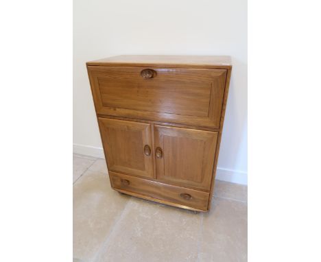 An Ercol blonde elm mid 20th Century cabinet with fall front to top over two cupboard doors with a shelf to the interior, abo