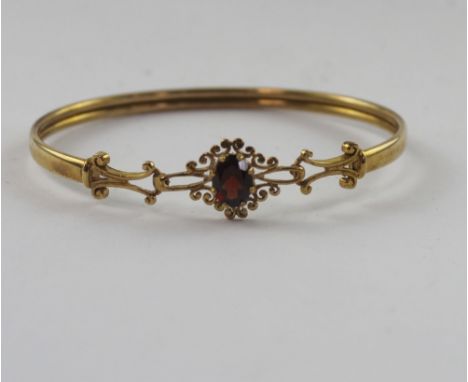 A 9ct yellow gold bangle with central section with an oval garnet in a floral scroll setting, approx 6.3g.
