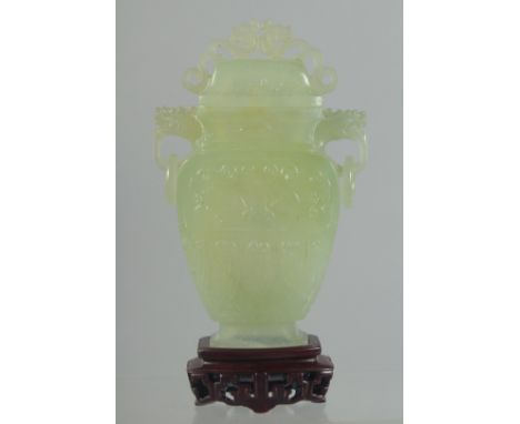 A CHINESE CARVED JADE LIDDED VASE AND WOODEN STAND, the vase with archaic style carving and drop ring handles, 18.5cm high (o