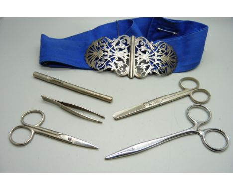 A silver nurse's belt buckle with silver nursing badge, scissors, a thermometer in metal tube and tweezers 
