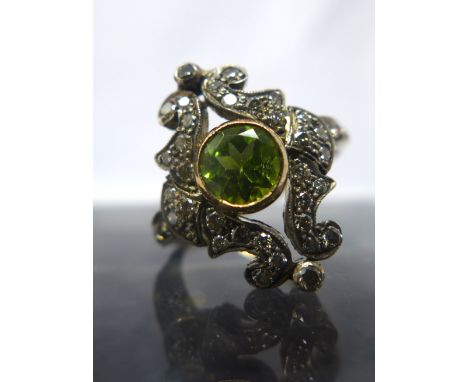 A Belle Epoque yellow gold, diamond and peridot ring, the central round bezel-set peridot framed by a scrolling surround of d
